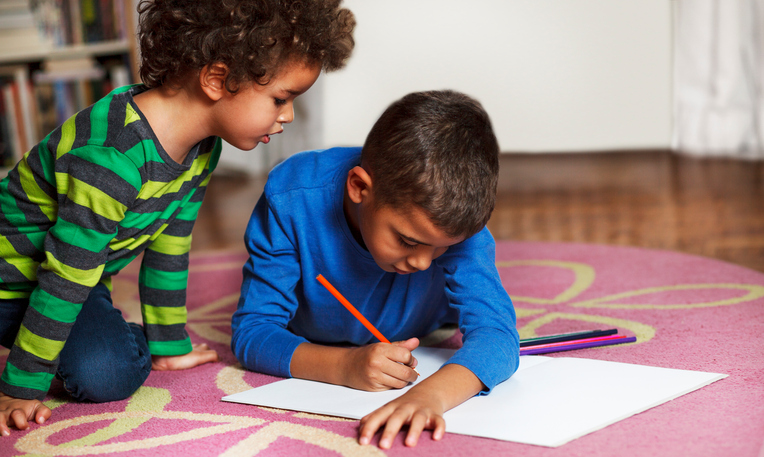 Cute school boys lying on the carpet, at home, drawing with crayons.