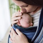 Close up, beautiful young mother kissing her newborn baby son in sling at home