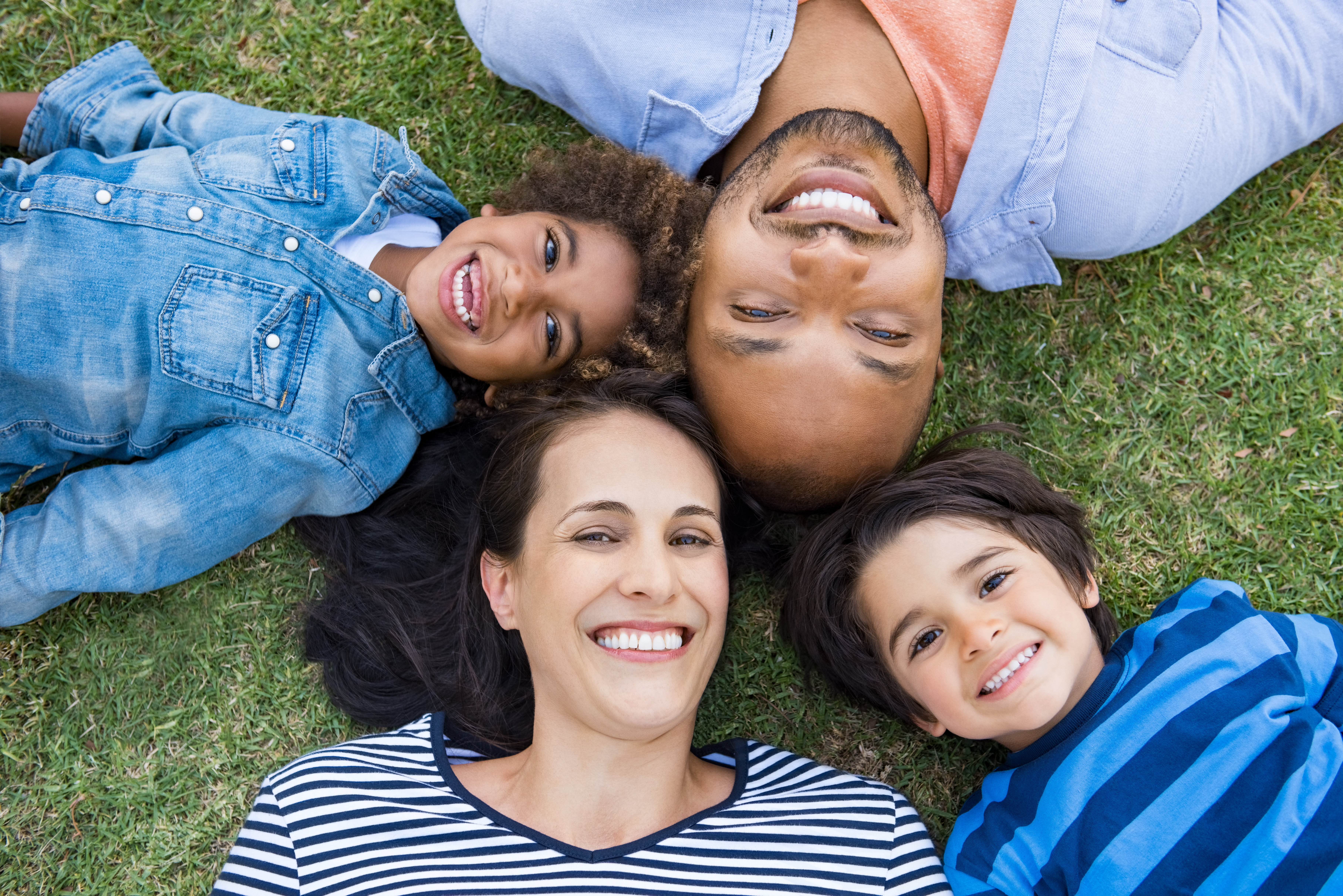 Overhead view of cheerful family lying on grass in a circle and looking at camera. High view of multiethnic family lying on grass at park. African father and hispanic mother with their smiling children